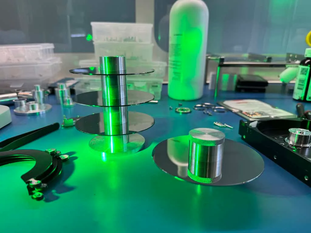 A stack of hard drive platters and head combs staged on a clean room workbench under green inspection light during a platter transplant